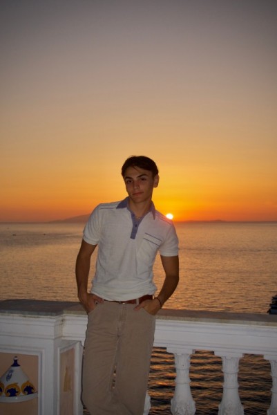 Photo shows a young, caucasian, male student with dark hair standing on a balcony with the ocean or a large lake behind him, with an orange, yellow, and gold sunset on the horizon. He is wearing a white t-shirt, khaki pants, and a brown belt.
