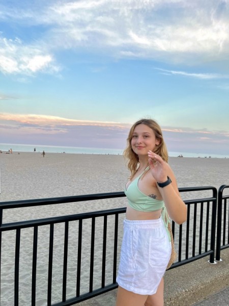 Photo shows a young, caucasian, female student standing at the beach with the sand and sky behind her. The sky is blue and purple. She is looking back over her left shoulder and is wearing a blue top and white shorts, has blonde hair, and has her hand raised in a waving position.