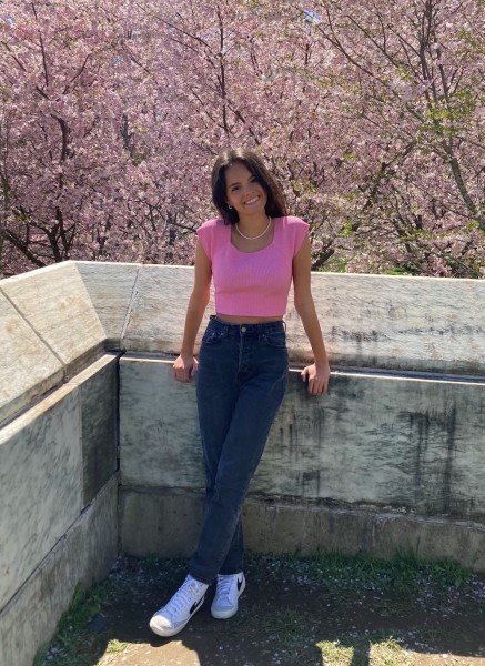Photo shows a young, caucasian, female student in a bright pink cropped shirt, jeans, and white sneakers. She is standing on a stone balcony in front of a pink cherry blossom tree.