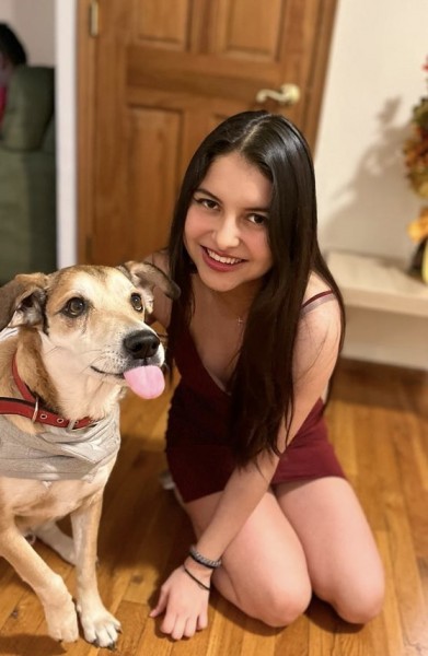 Photo shows a young, Hispanic, female student kneeling down next to a brown dog in a foyer or living room. The student is wearing a spaghetti-strap red dress and has long dark hair and brown eyes. The dog has its tongue sticking out.