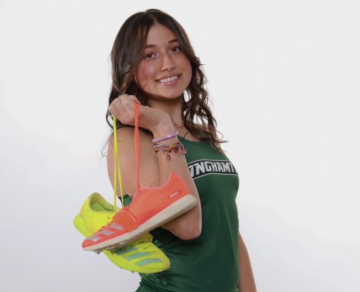 Photo shows a young, caucasian, female student posing while holding sneakers in a green Binghamton University track and field jersey. She has brown hair and is holding running shoes.