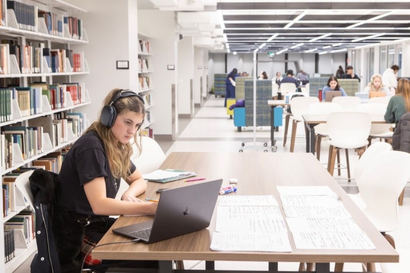 A student wearing headphones sits at a table in the library, studying.