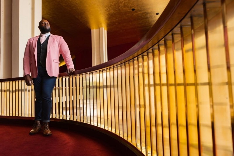 Thsombe Selby stands at the top of stairs on a red carpeted balcony, looking off to the side