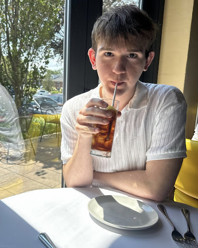 Photo shows a young, caucasian, male student sitting at a table at a restaurant, holding a drink with the straw in his mouth as he drinks. He is sitting next to a window and is wearing a white shirt.