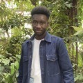 Portrait of a young, African American, male student. Student is dressed in a blue button-up long-sleeved shirt with a white shirt underneath and is standing among many plants in a greenhouse.