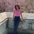 Photo shows a young, caucasian, female student in a bright pink cropped shirt, jeans, and white sneakers. She is standing on a stone balcony in front of a pink cherry blossom tree.