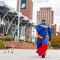 A robot dressed like Superman walks with a robot dog on campus.