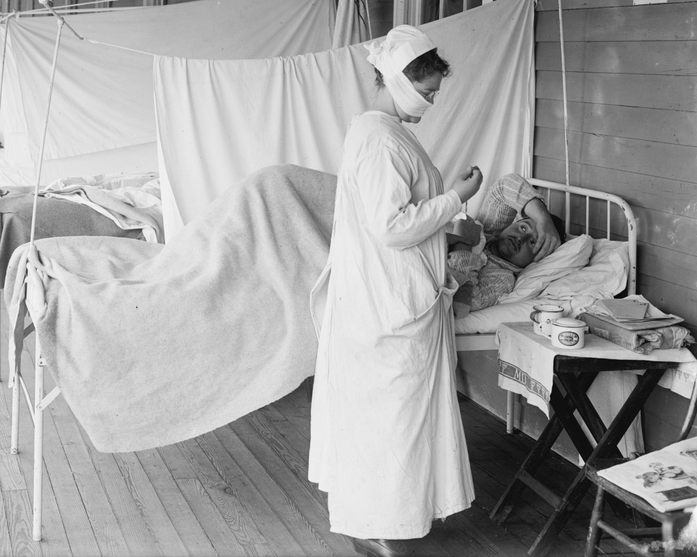 Photo of Walter Reed Hospital, Washington, D.C., during the great influenza pandemic of 1918–1920. Patients are set up in rows of beds on an open gallery, separated by hung sheets. A nurse wears a cloth mask over her nose and mouth.