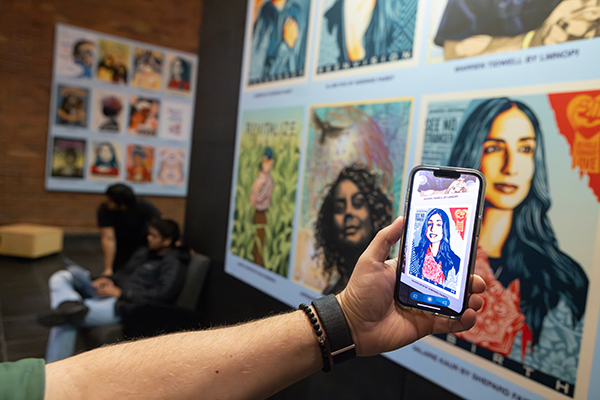 A Glenn G. Bartle Library visitor holds a phone toward a collection of art portraits displayed on the first floor of the library, showcasing the Augmented Reality app that animates the image.