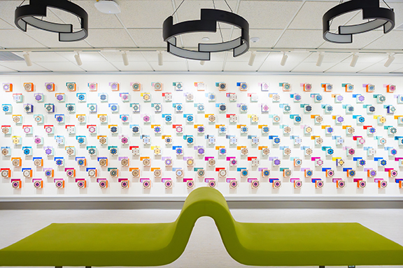 The installation on the third floor of Glenn G. Bartle Library gallery is a data-driven tribute to the collection of Binghamton University Libraries. A lime green curved bench sits in the foreground, as 3D blocks with different colors and patterns are seen on a white wall.