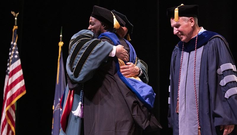 Randall Eduouard, assistant vice president for student affairs and dean of students, gets a hug from President Harvey Stenger after being hooded for earning his Doctor of Education. Provost Donald Nieman looks on.