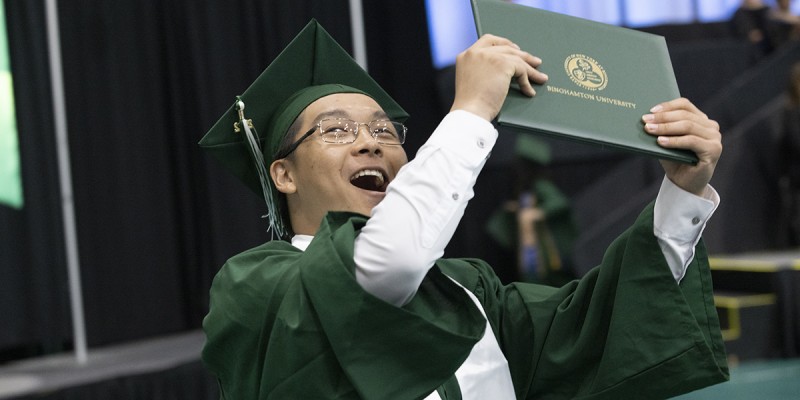 A newly minted graduate of the School of Management shows off his diploma (cover) to family in the stands after crossing the stage at the Events Center.