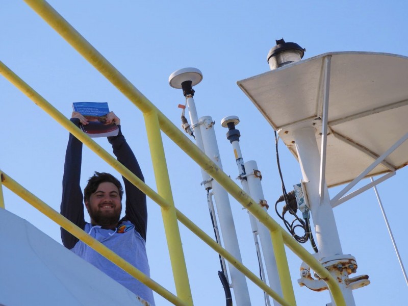 Michael Kashinsky holds his geology textbook aloft on the deck of the JOIDES Resolution, a deep-sea research vessel.