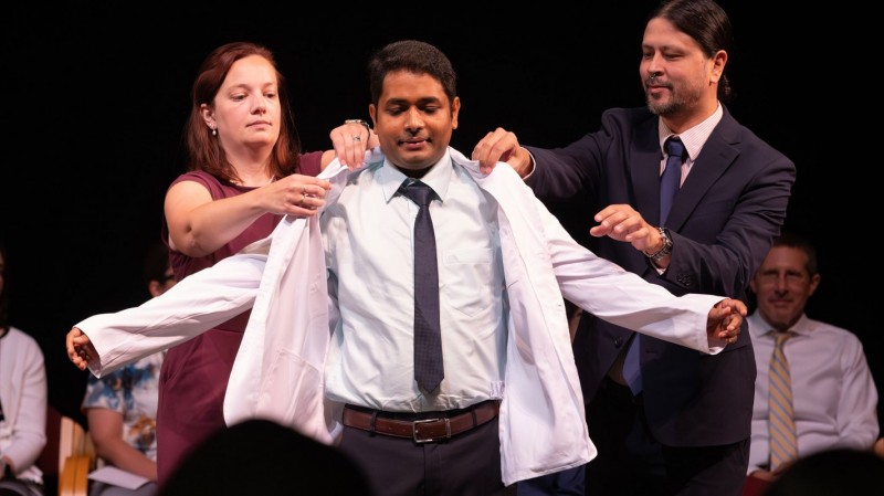 Clinical Associate Professor Erin Pauling (left) and Clinical Assistant Professor Anthony Hopkins (right) help P1 student Julfikar Ali put on his white coat.