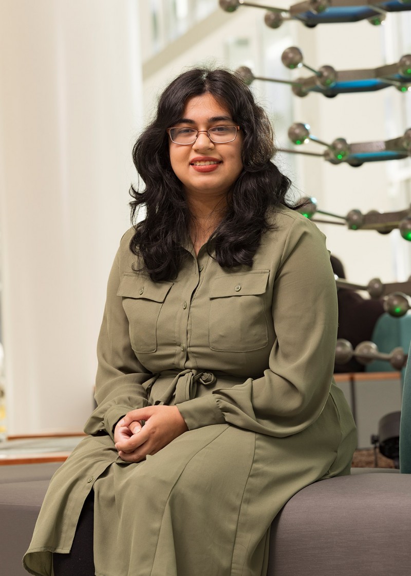 Lamya Islam sitting near the sculpture of a caffeine molecule in the School of Pharmacy's atrium.