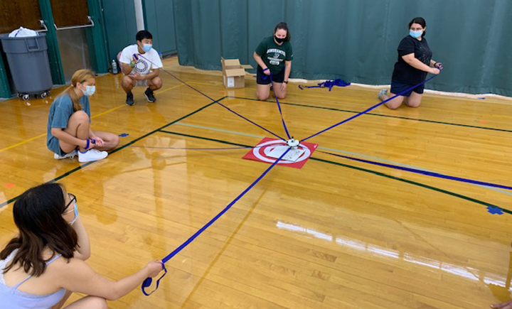 Students in a Healthy College Living class play a socially distanced transfer game. The students must maneuver the straps to pick things up and get them into the bucket in the middle.