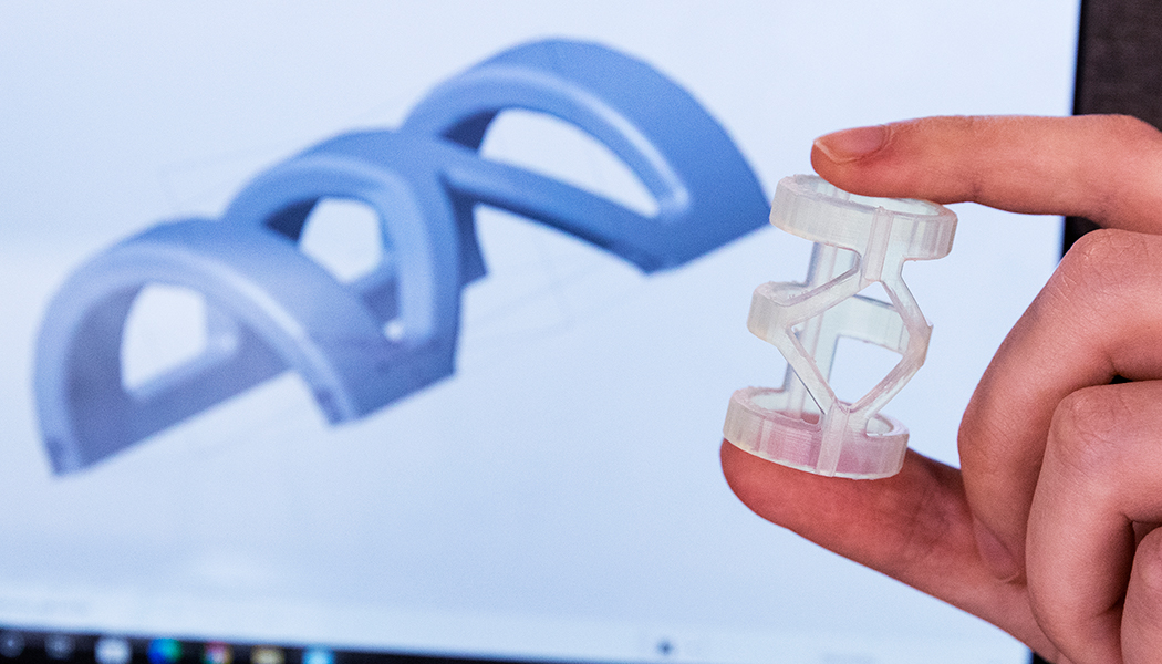 The blue image is a computer-aided design (CAD) model of the orthosis the students developed for people with injuries to the proximal interphalangeal joint (PIPj) of the fourth digit of the hand. In the foreground is a 3D-printed model of the device.