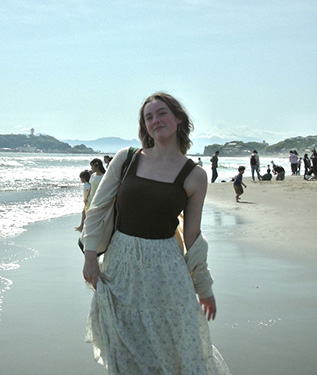 A woman with a brown tank top, sweatshirt with one of her shoulders uncovered, stands on the edge of water with small waves visible in the background