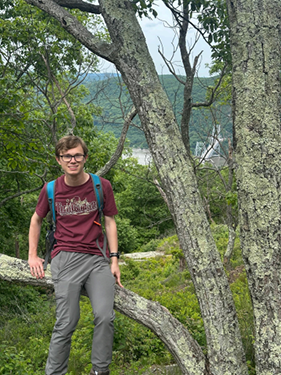 A man wearing glases and a blue backpack sits on the branch of a tree in a heavily forested area