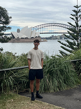 A man wearing shorts and a t-shirt with his hands in his pocket stands in front of some green bushes with the Sydney Opera House in the background