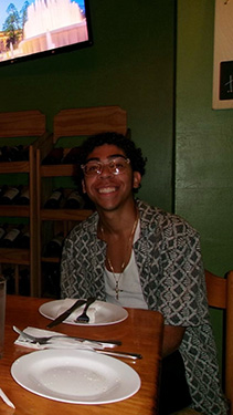 A man wearing glasses and an open-front shirt seated at corner of a table in a restaurant