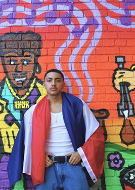 A man with a white shirt standing in front of a mural with a flag over his shoulder