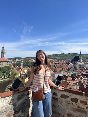 A woman with a pink and white striped tank top standing near a wall with the roofs of buildings in the background on a sunny day