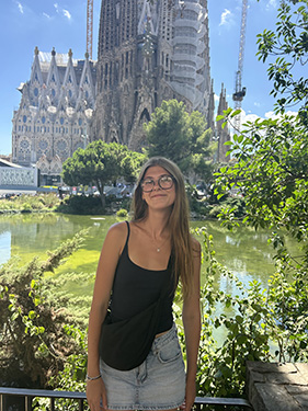 A woman wearing a black tank top and glasses standing in front of a pond with a large church in the background