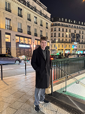 A man wearing a black coat with his hands in his pocket at the top of a staircase leading downward into an underground train at night