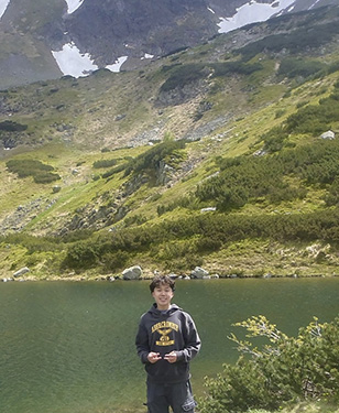 A man in a hooded sweatshirt stands in front of a small lake with green hilly mountains in the background