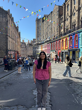 A woman in a pink shirt and grey jeans stands in a plaza whose doors are painted bright colors on the first level with pennant flags streching between buildings