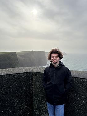 A man wearing a black jacket and blue jeans poses at the Cliffs of Moher in ireland on a semi-cloudy day