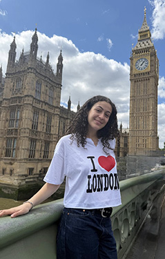 A woman wearing a white t-shirt that says I love London stands on a bridge with Big Ben and Parliament in the background