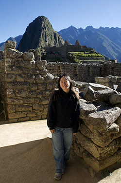 A woman in a black hooded sweatshirt and blue jeans stands in the ruins of Machu Pichu during the day