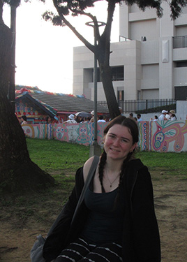 A woman wearing a black tank top sits on the ground in front of a grey wall containing grafitti