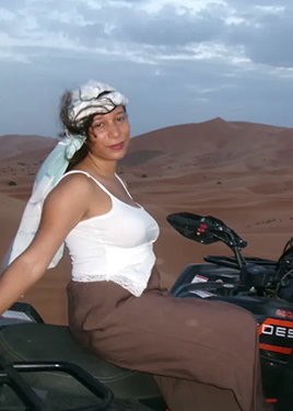 A woman with white shirt and a brown skirt sits on an off-road vehicle in the desert on a cloudy day