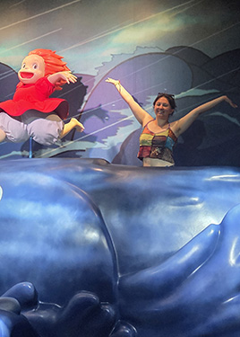 A woman stands with both of her hands up and to the side of her head next to a puppet with red hair who is running across crashing waves
