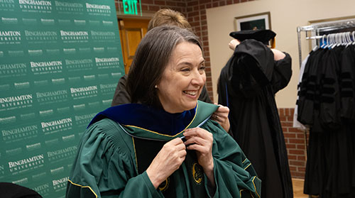 Anne putting on cap and gown in front of binghamton background
