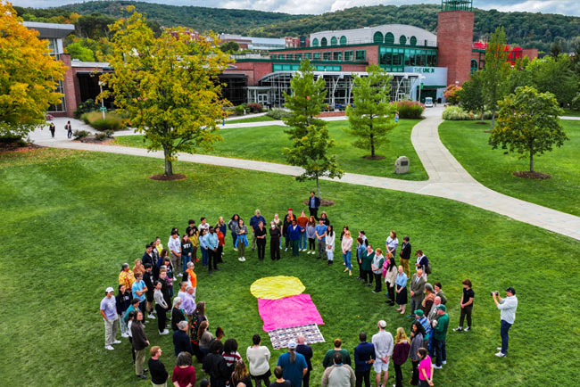 A group gathers in a circle in the Peace Quad lawn with buildings and trees in the background.