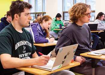 Students attending a classroom lecture while taking notes and working on laptops.
