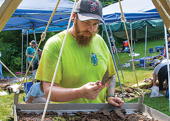 Student participating in an archaeological field excavation while examining artifacts at a screening station.