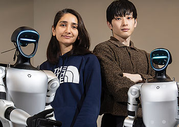 Two Binghamton University students standing with humanoid robots in a robotics laboratory.