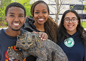 Three Binghamton University students smiling beside the Bearcat statue on campus.