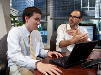A male student sits in an office looking at a computer with an adult male who is explaining something photo