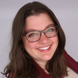 Professional Headshot of Meghan Crist. A white woman with brown hair and glasses, in a red blazer and a beige turtleneck. She's smiling.