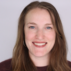 Professional headshot of Shira Clark. A white woman, with brown wavy hair, in a dark red sweater. She's smiling.