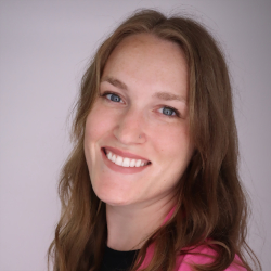Professional headshot of Shira Clark. A white woman, with brown wavy hair, in a pink blazer and a black shirt. She's smiling.